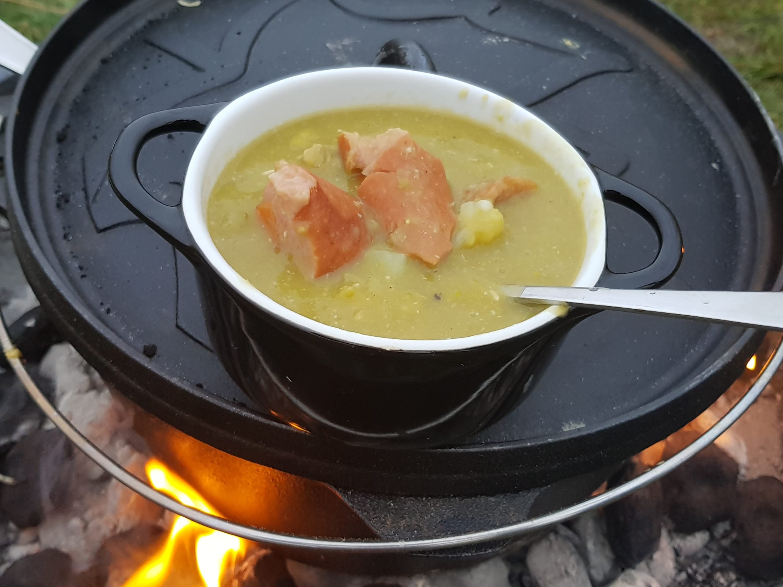 Petromax Dutch Oven gietijzeren stoofpan op open vuur met een kom erwtensoep en rookworst op het omgedraaide deksel