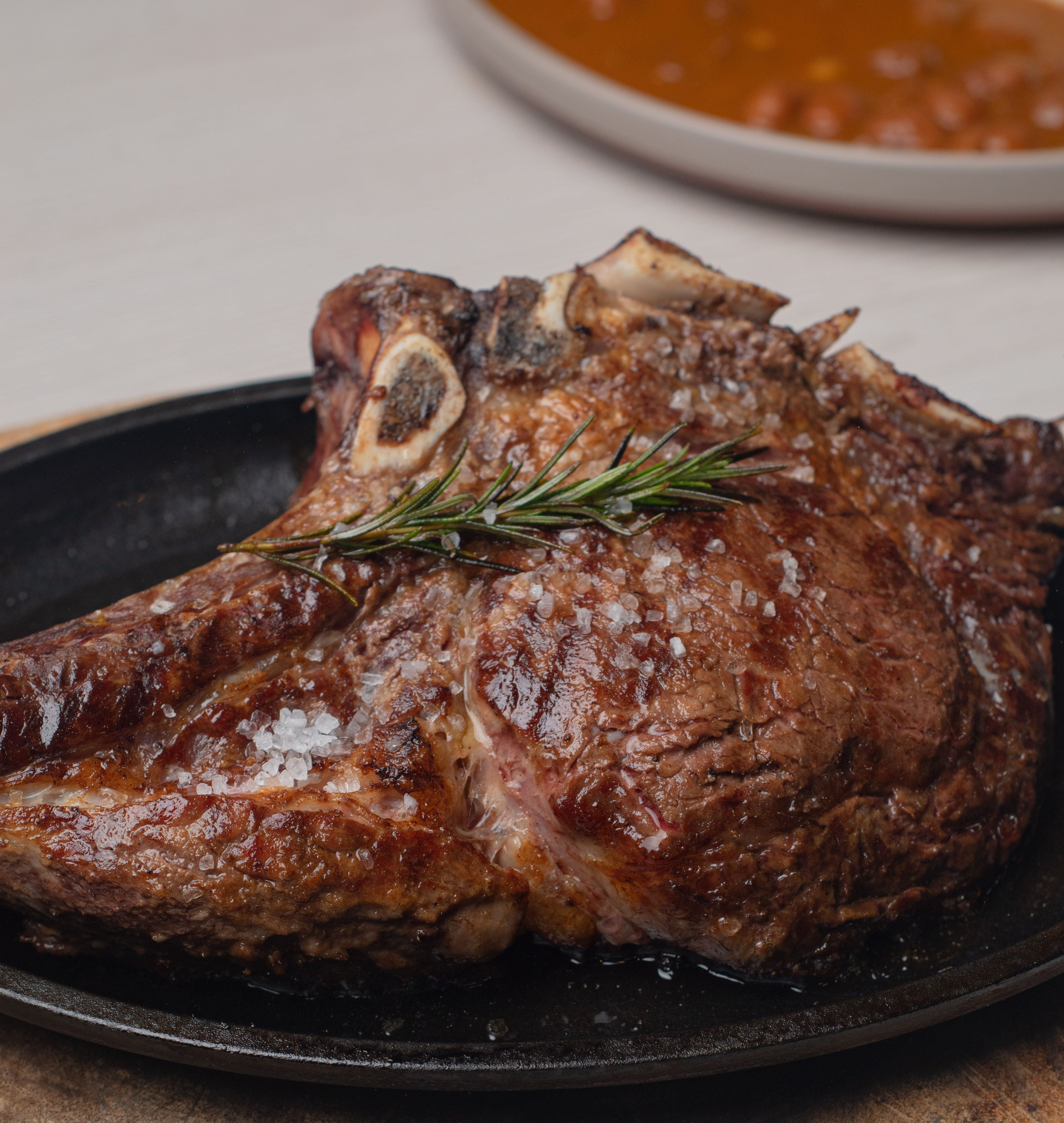 Een grote, gegrilde T-bone of côte de boeuf steak met grof zeezout en een takje rozemarijn op een zwarte gietijzeren schaal.