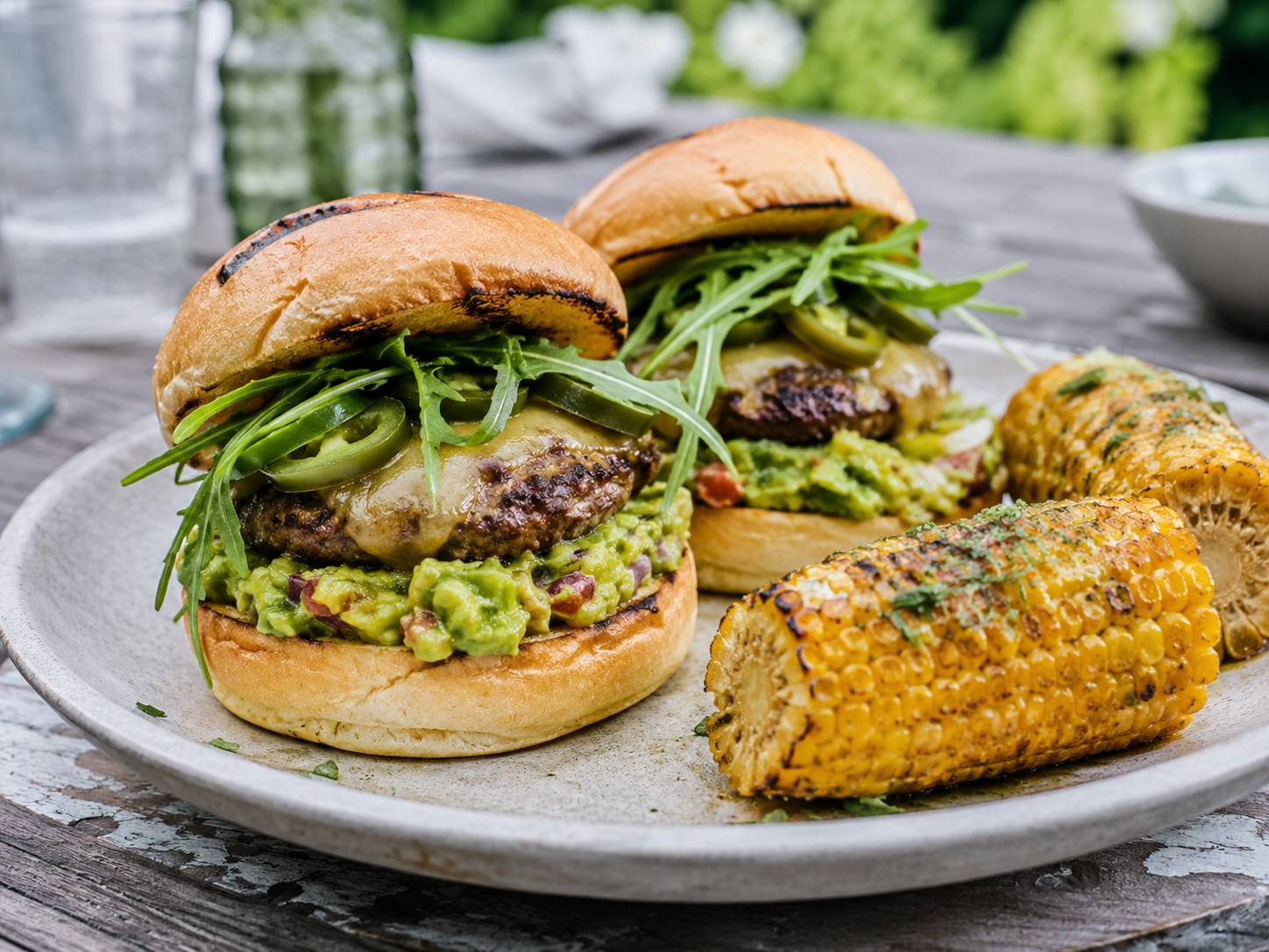 Een smakelijke foto van twee gegrilde burgers op een bord, belegd met guacamole, gesmolten kaas, jalapeños en rucola. Ernaast liggen twee geroosterde maïskolven bestrooid met kruiden. Het bord staat op een rustieke houten buitentafel met een onscherpe, groene achtergrond.