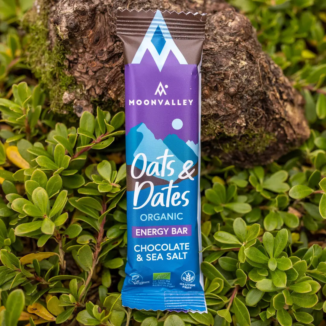 Moonvalley Oats and Dates Chocolate and Sea Salt veganistische energy bar in paarse verpakking tegen een achtergrond van mos en boomstam