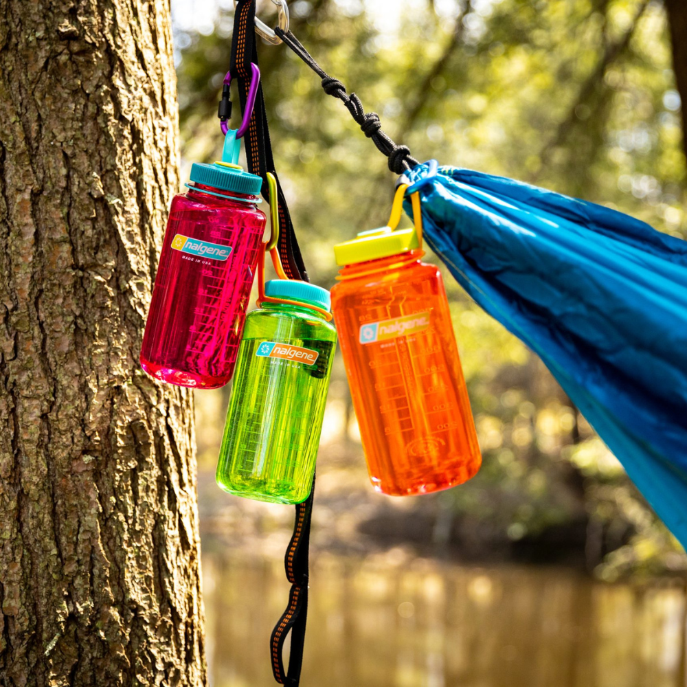 Drie gekleurde Nalgene waterflessen in rood, groen en oranje hangend aan een zwarte band bij een boom en blauwe hangmat